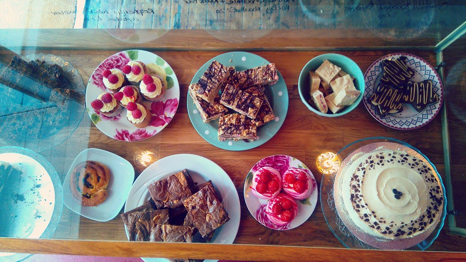 Vintage floral plates seen through the counter glass: raspberry cupcakes, flapjacks, fudge, meringues, brownies and a cheesecake.