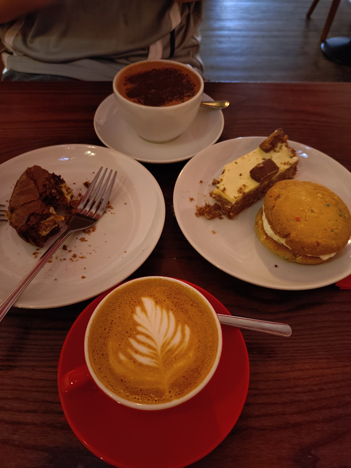 A flat white on a red saucer, a hot chocolate, a brownie, a caramel traybake slice and a sprinkle cookie sandwich.