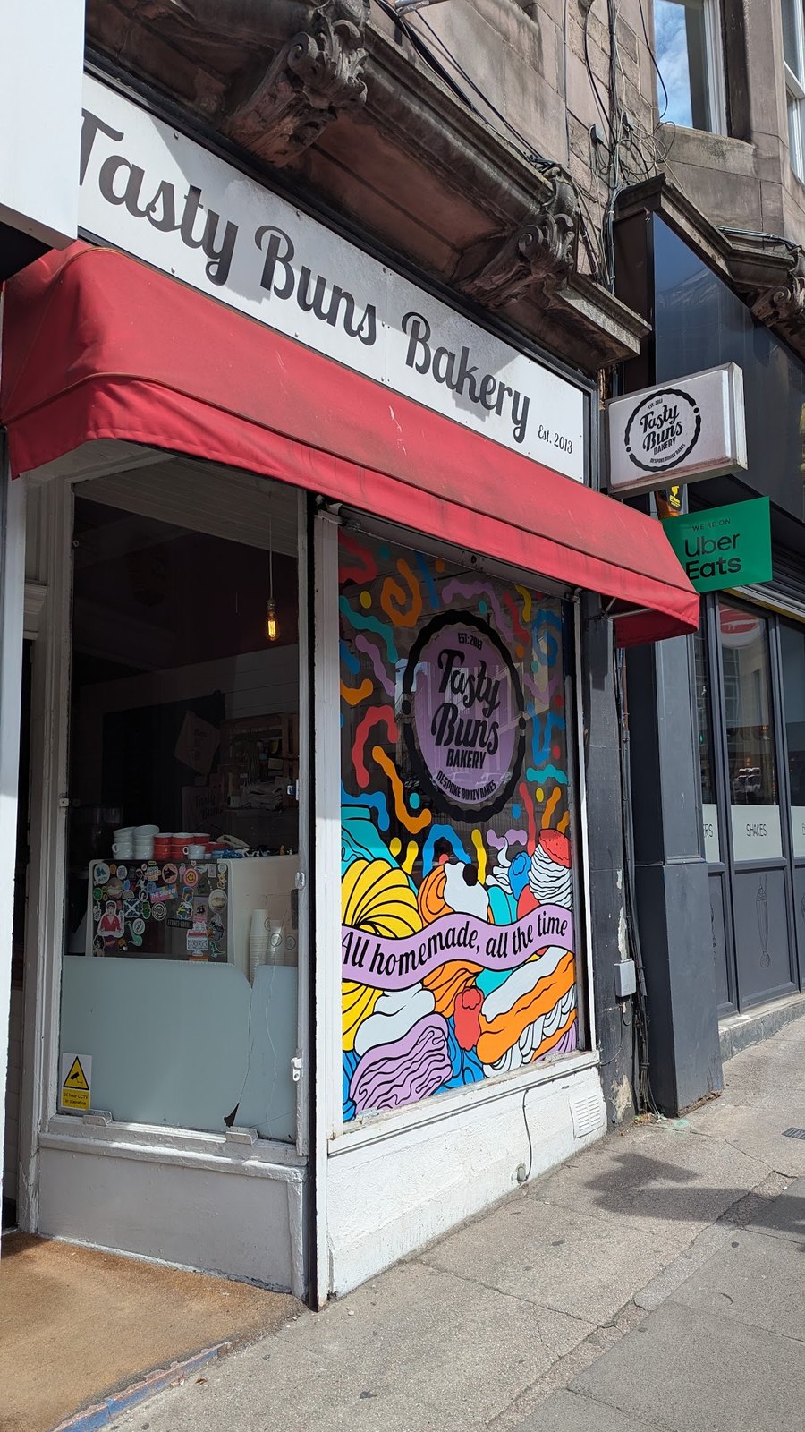 The shopfront: Tasty Buns Bakery in script on a white fascia with a red awning, and a bright mural reading All homemade, all the time.
